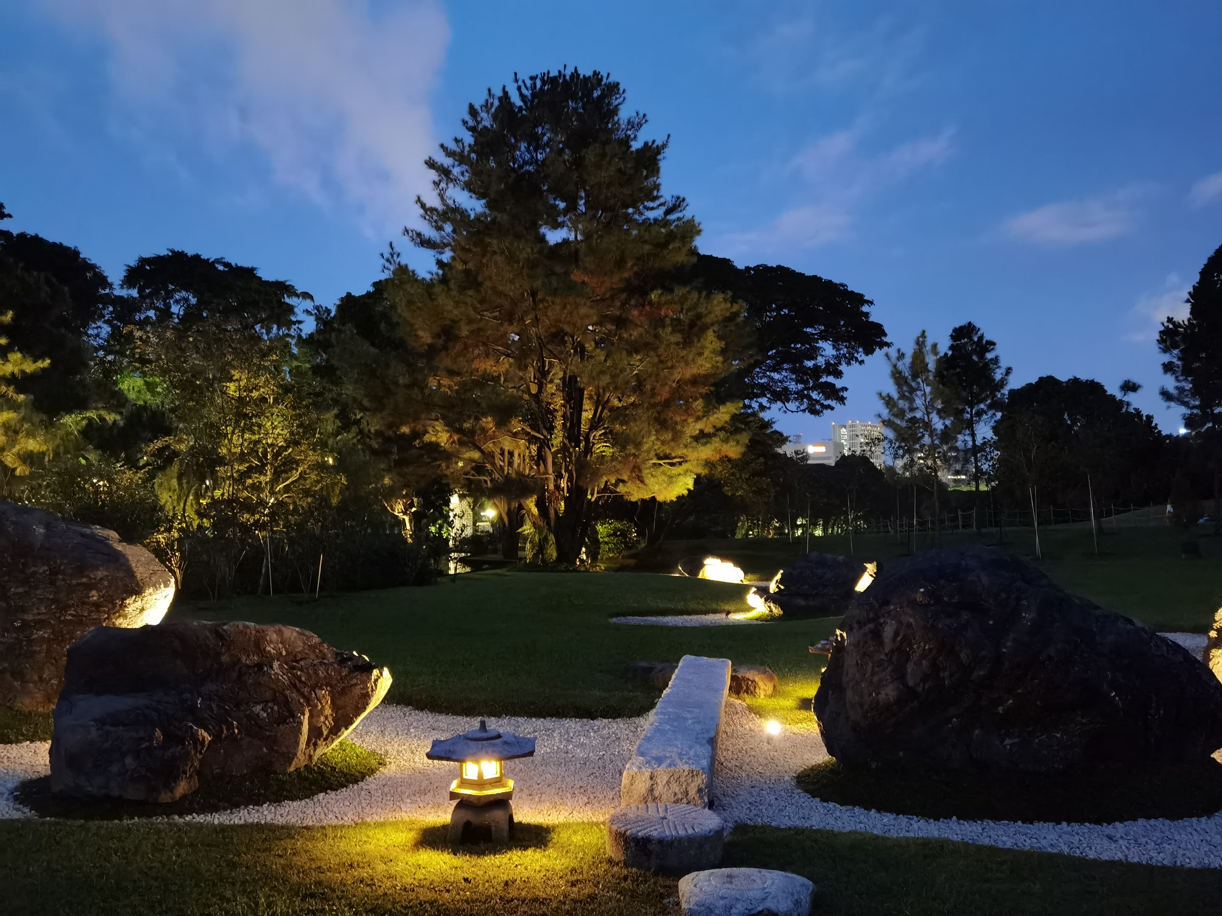 Japanese garden at night with trees, rocks, gravel paths, stone lanterns, and building in background.